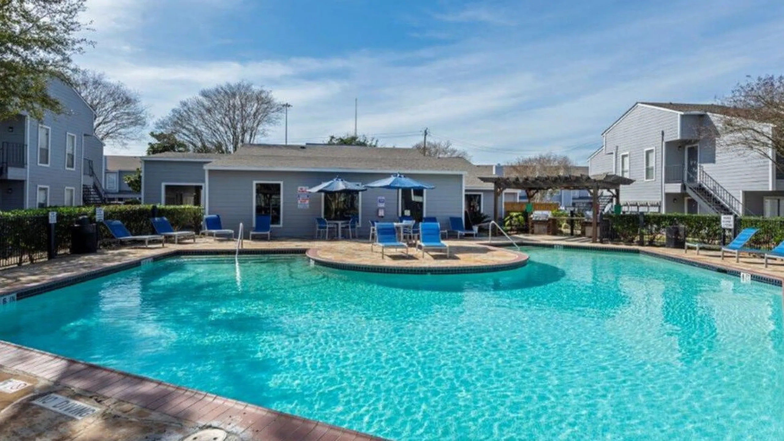 Inviting outdoor pool area with sun loungers, umbrellas, and surrounding modern apartments basking under a vibrant blue sky.