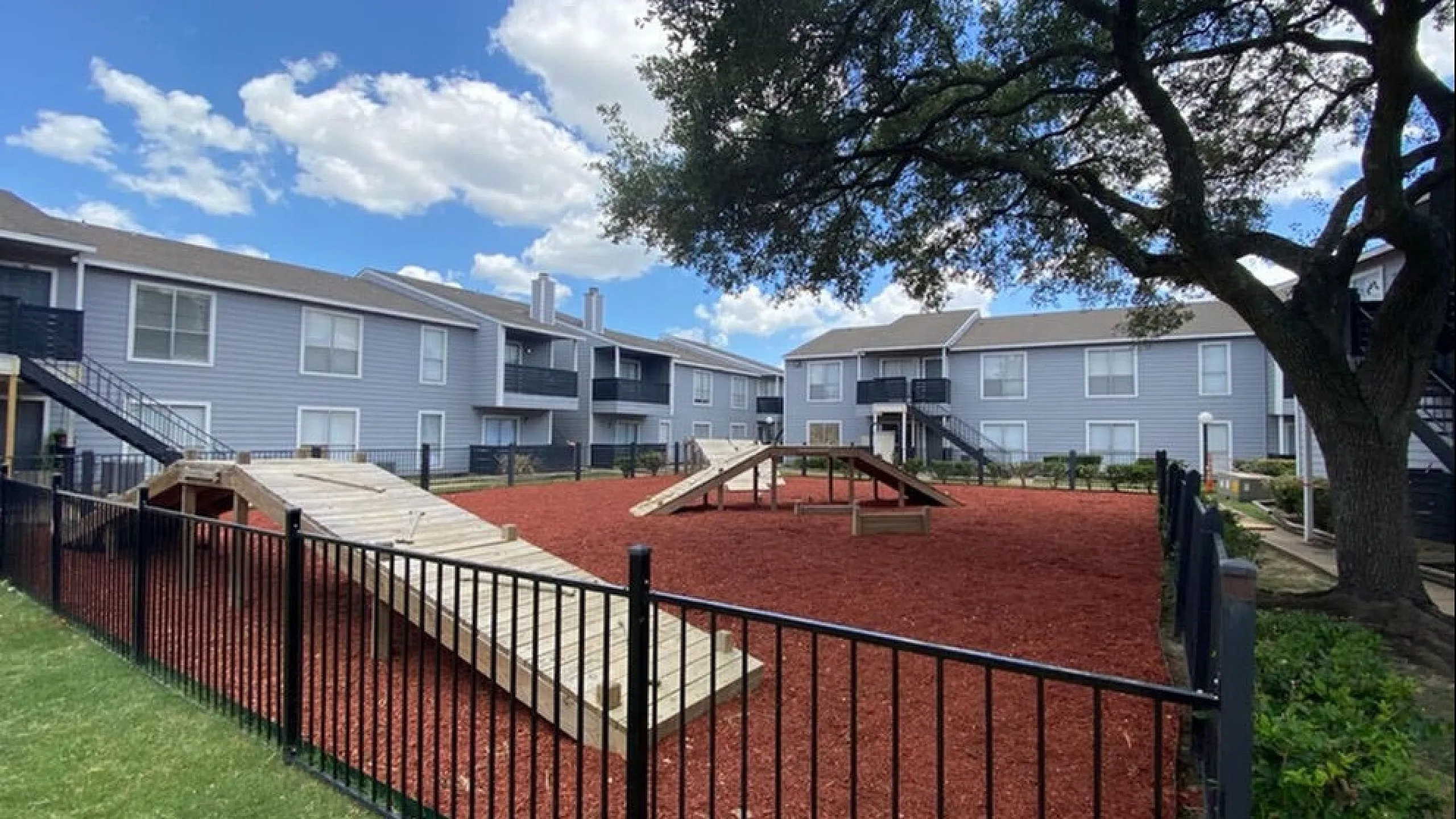 Charming apartment complex with stylish architecture, lush greenery, and fenced recreational area under a clear blue sky.