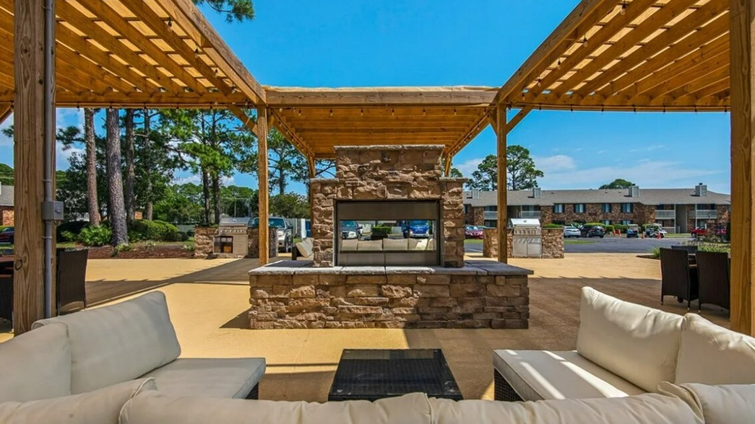 Chic outdoor seating area with a stone fireplace and elegant wooden pergolas, perfect for relaxation and entertaining under the blue sky.