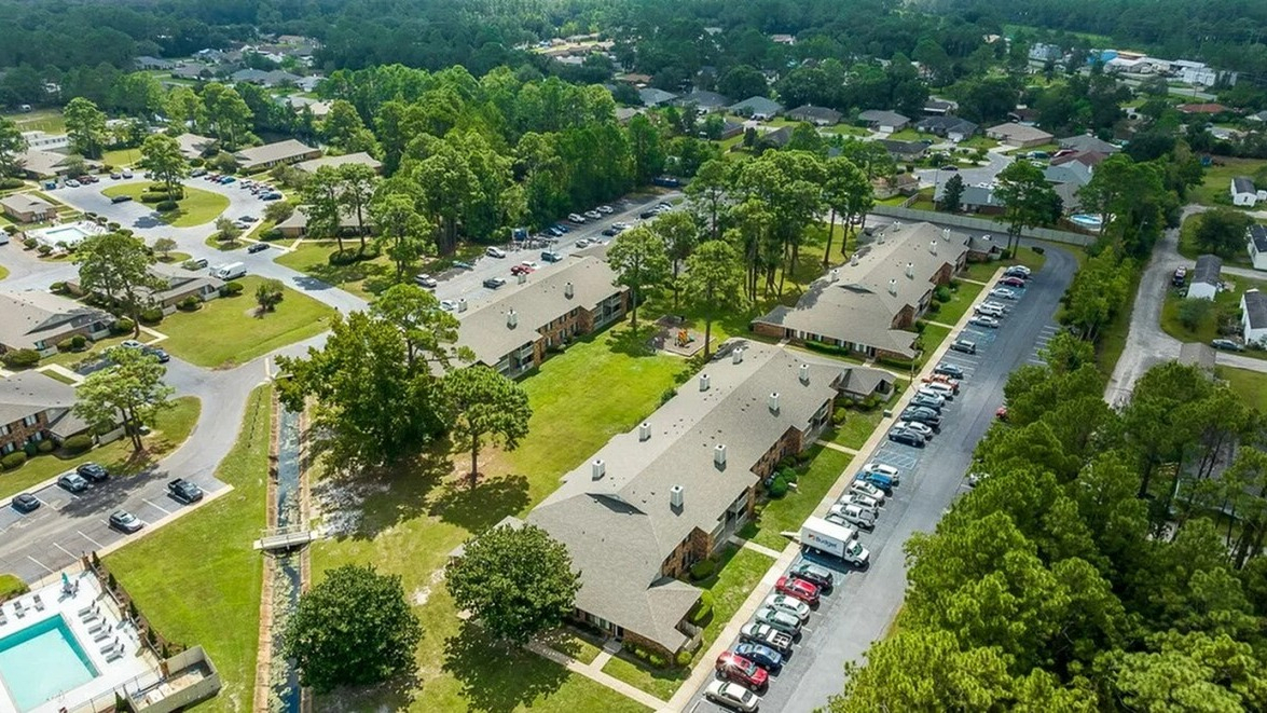 Aerial view of a charming apartment complex surrounded by lush greenery, featuring spacious parking areas and a refreshing outdoor pool for relaxation.