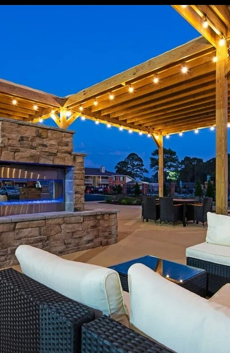 Charming outdoor lounge with cozy seating, stone fireplace, and ambient string lights under a wooden pergola.