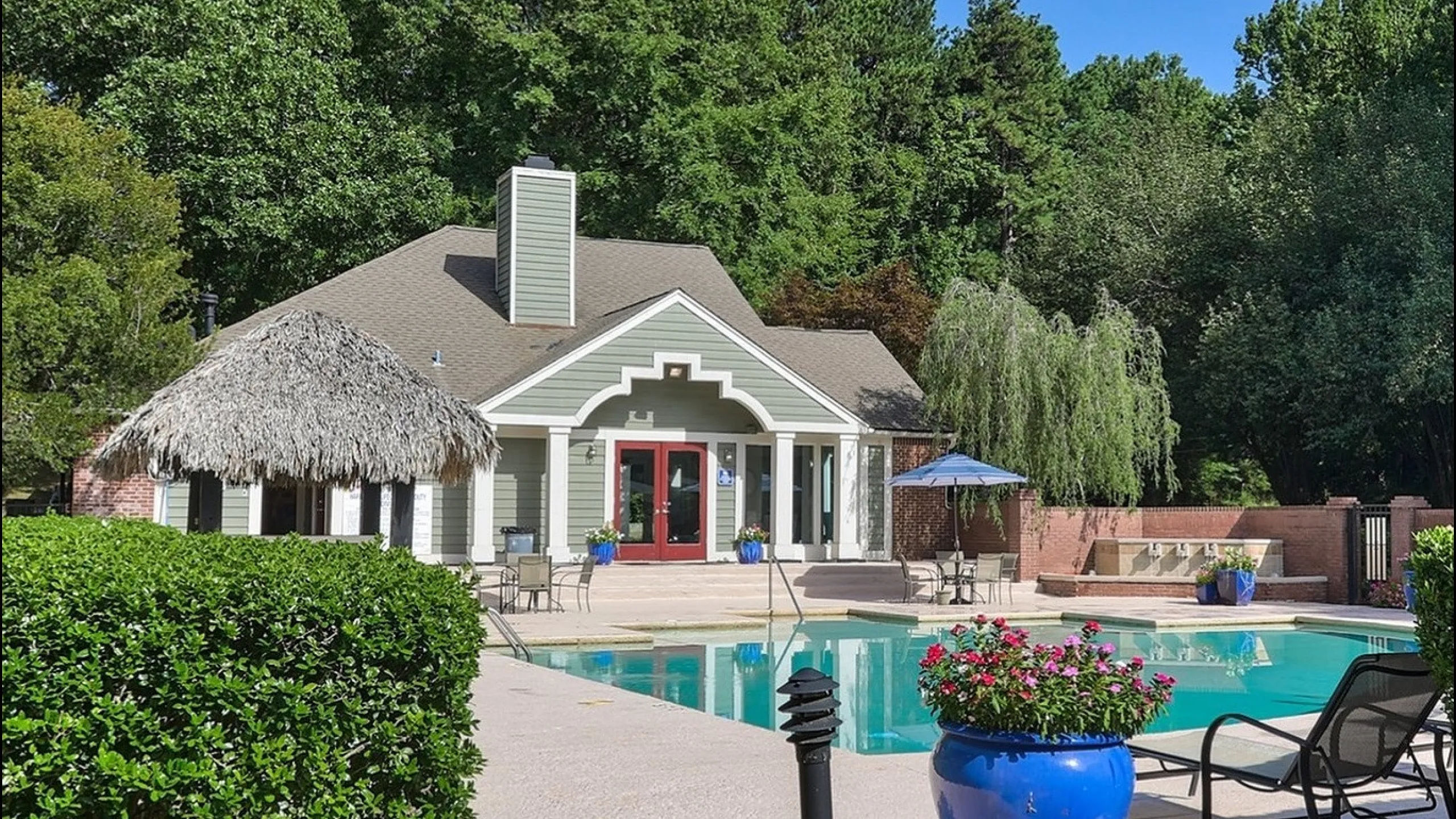 Charming community pool area with a thatched-roof cabana, surrounded by lush greenery and a stylish clubhouse.