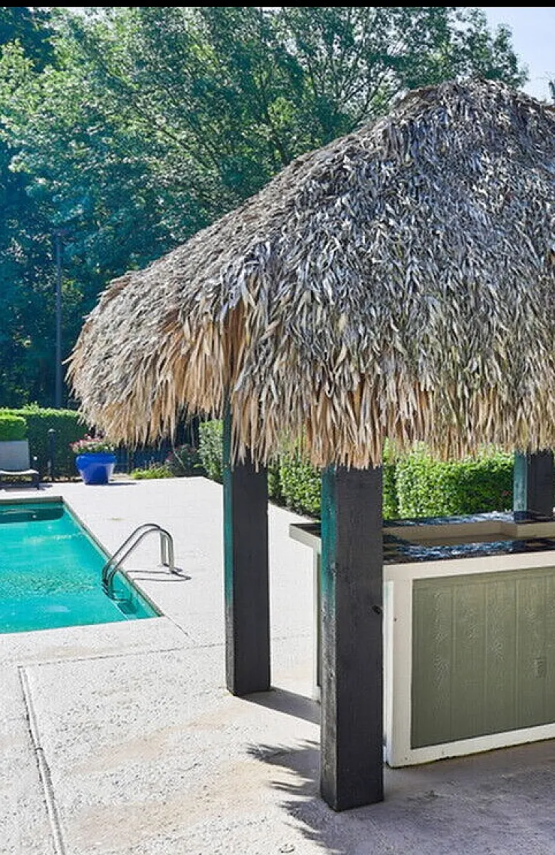 Inviting outdoor area with a charming thatched-roof tiki bar next to a sparkling swimming pool, surrounded by lush greenery and equipped with modern barbecue grills.