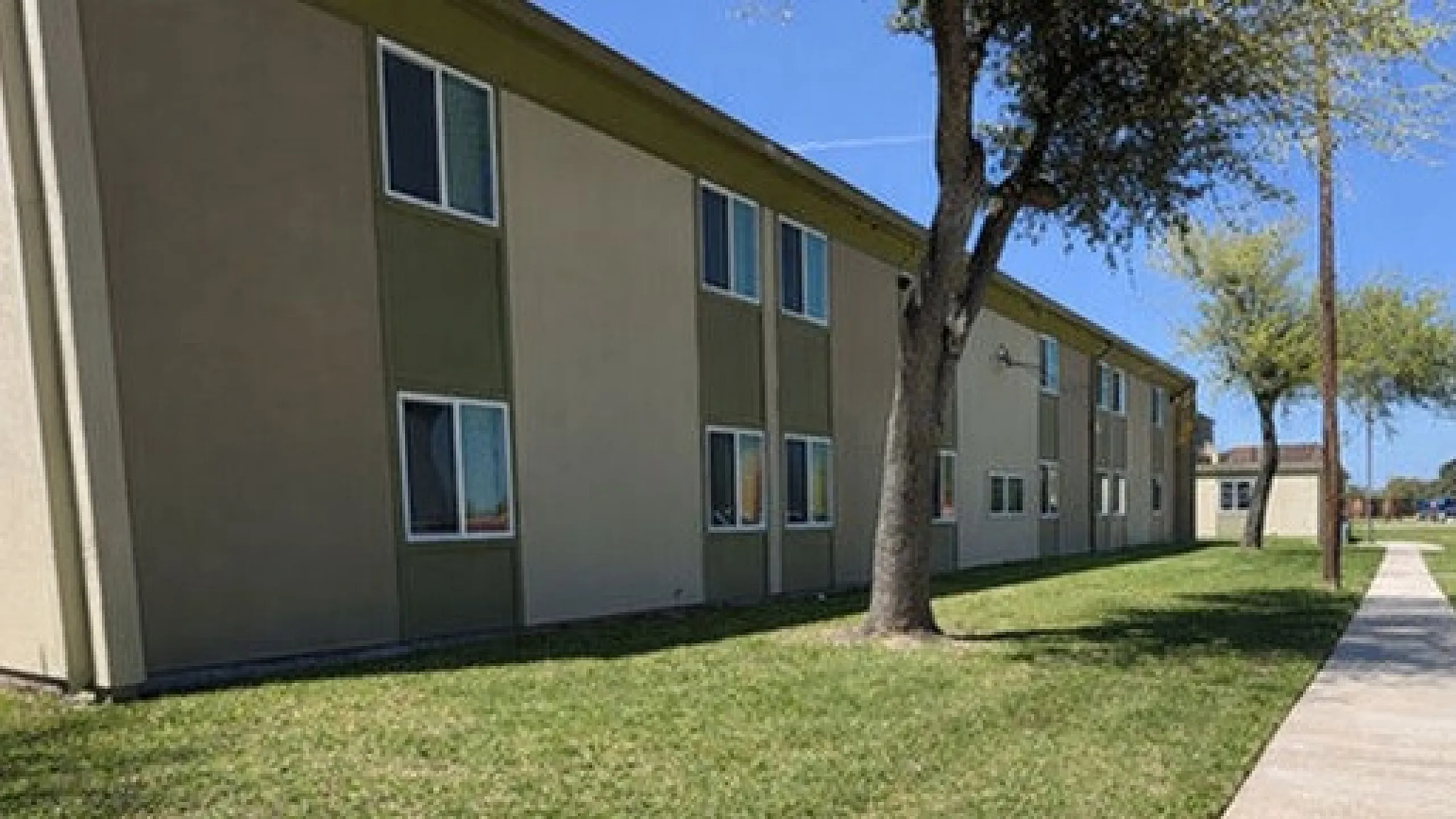 Charming apartment complex with well-maintained lawns, mature trees, and a spacious walkway perfect for strolling under sunny skies.