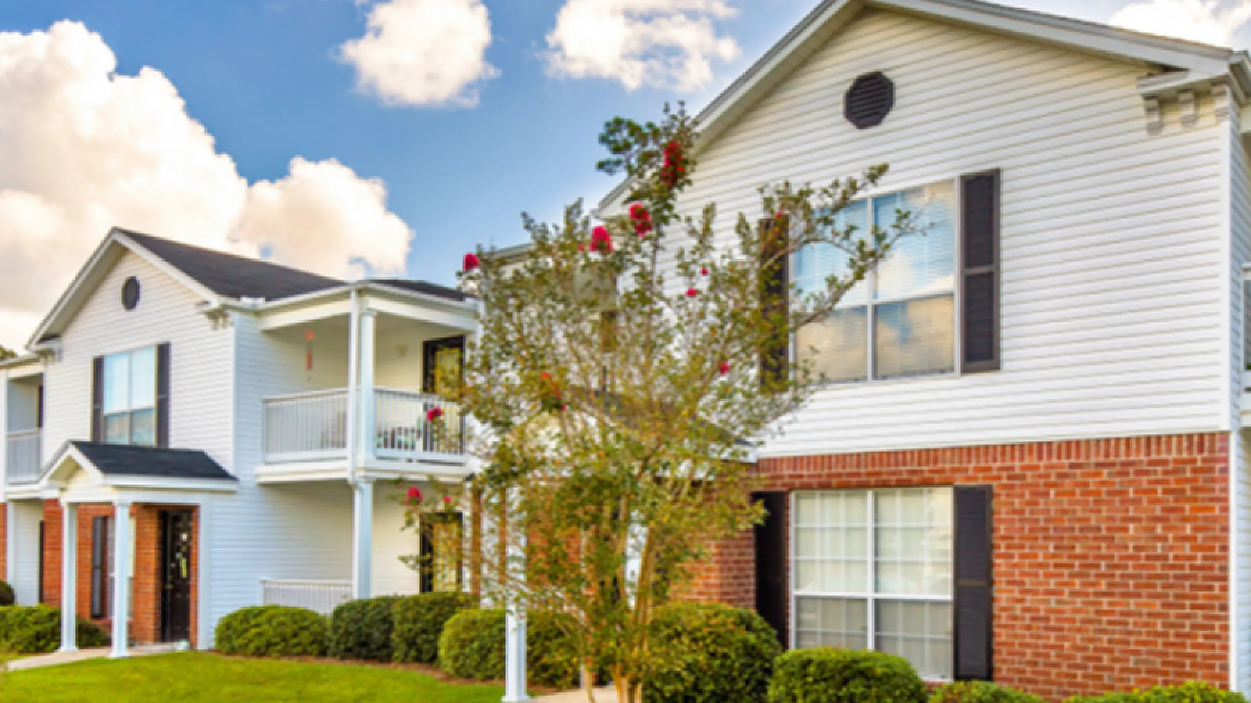 Charming two-story apartment complex with elegant white siding and classic brick accents, featuring inviting balconies and lush green landscaping under a bright blue sky.