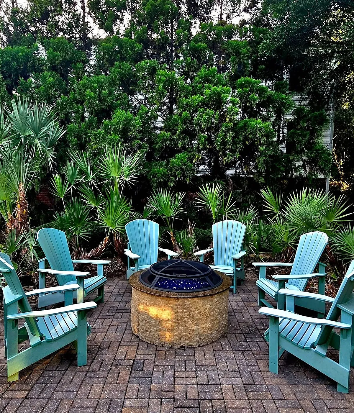 Inviting outdoor patio with turquoise Adirondack chairs around a cozy fire pit, surrounded by lush green foliage.