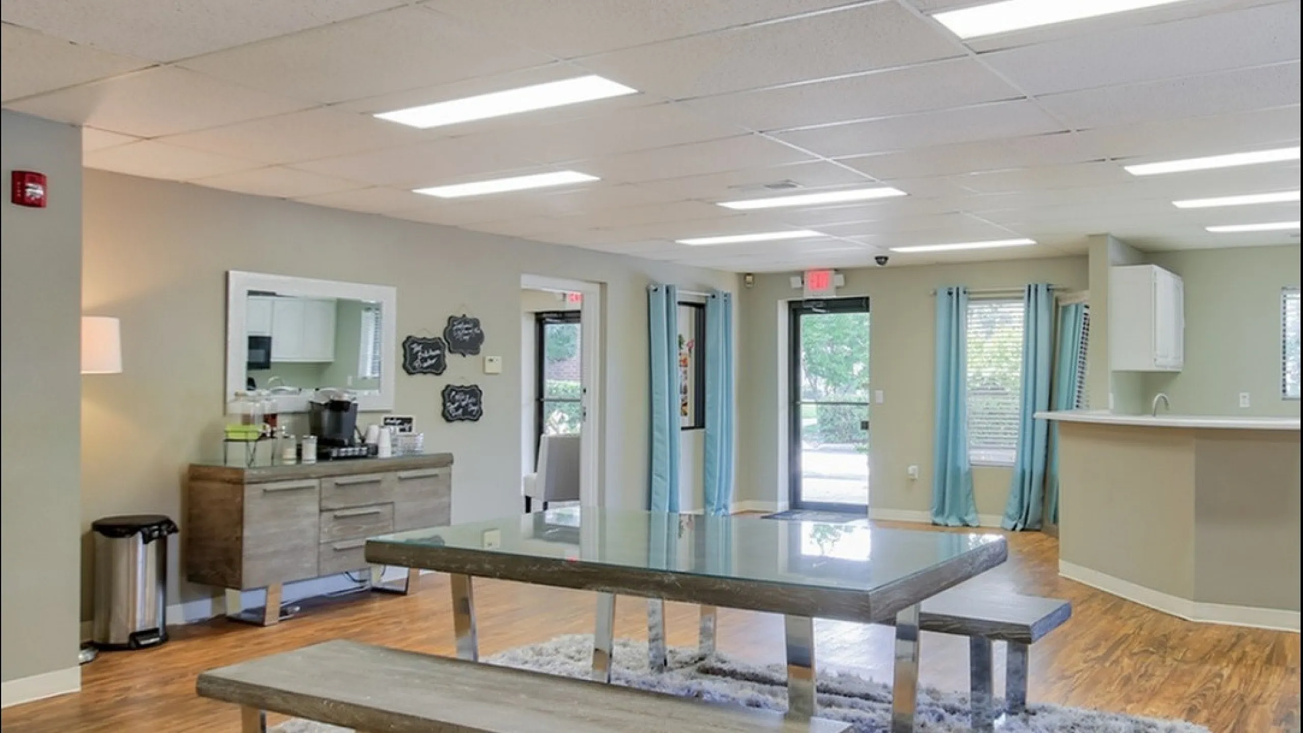 Modern communal room with sleek table and benches on plush rug, ample lighting, and a cozy coffee station.