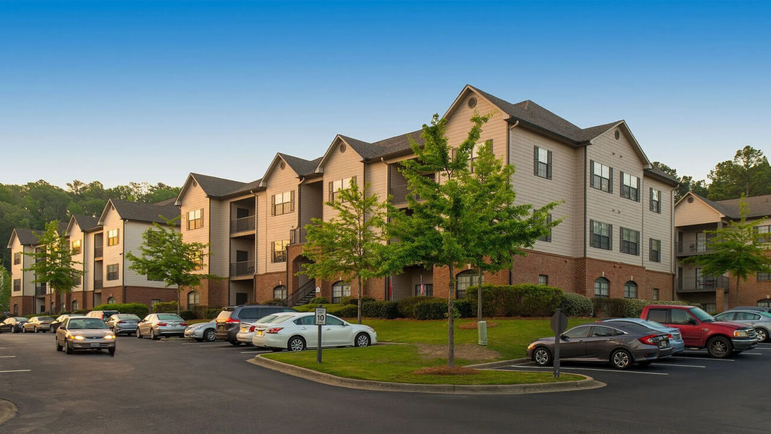 Charming apartment complex with modern architecture, ample parking, and lush greenery, set against a serene blue sky.