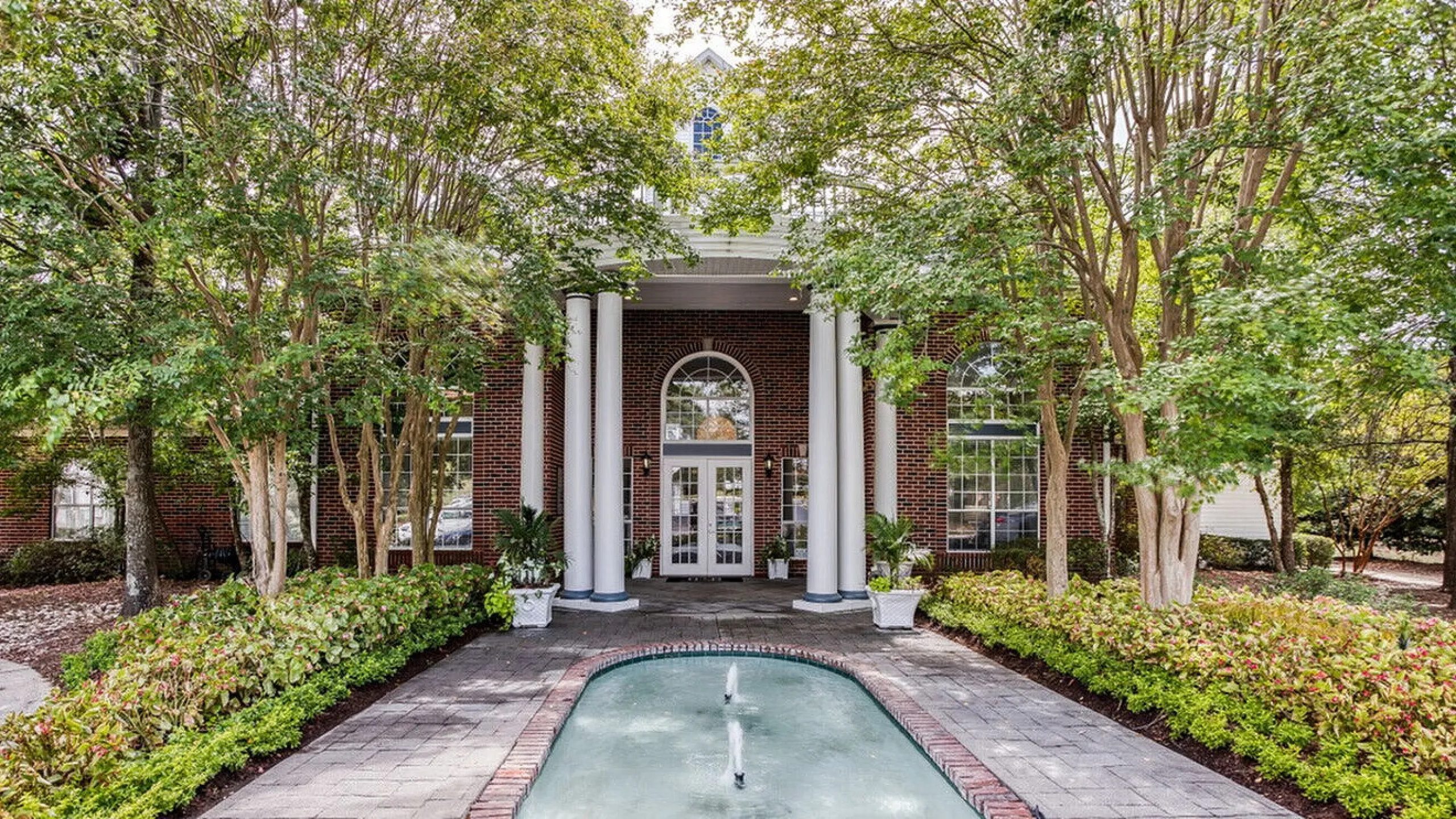Elegant entrance to a classic brick building with tall white columns, surrounded by lush greenery and a serene water feature.