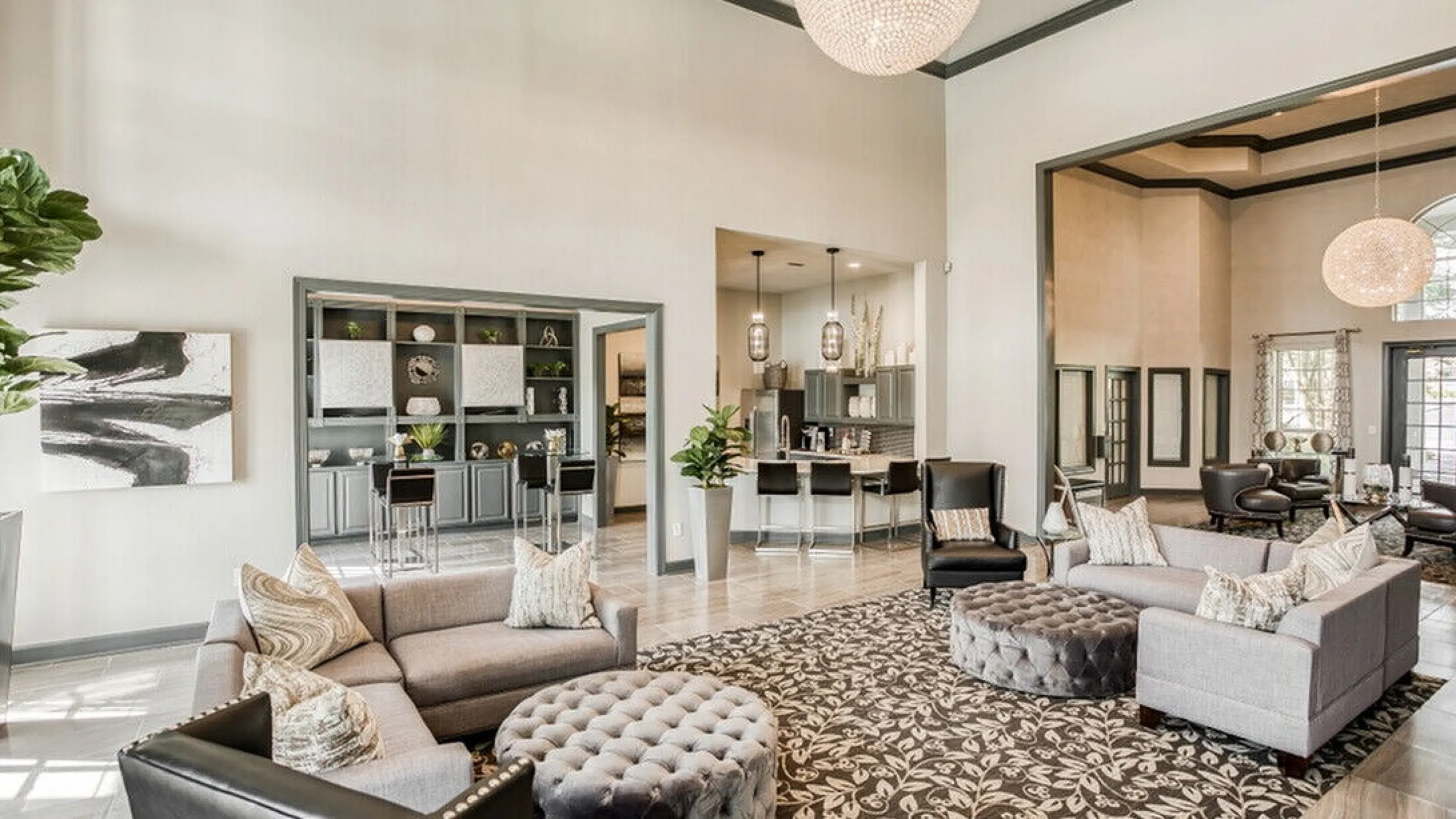 Elegant open living space featuring modern furnishings, plush seating, and a grand chandelier, leading to a stylish kitchen with bar stools.