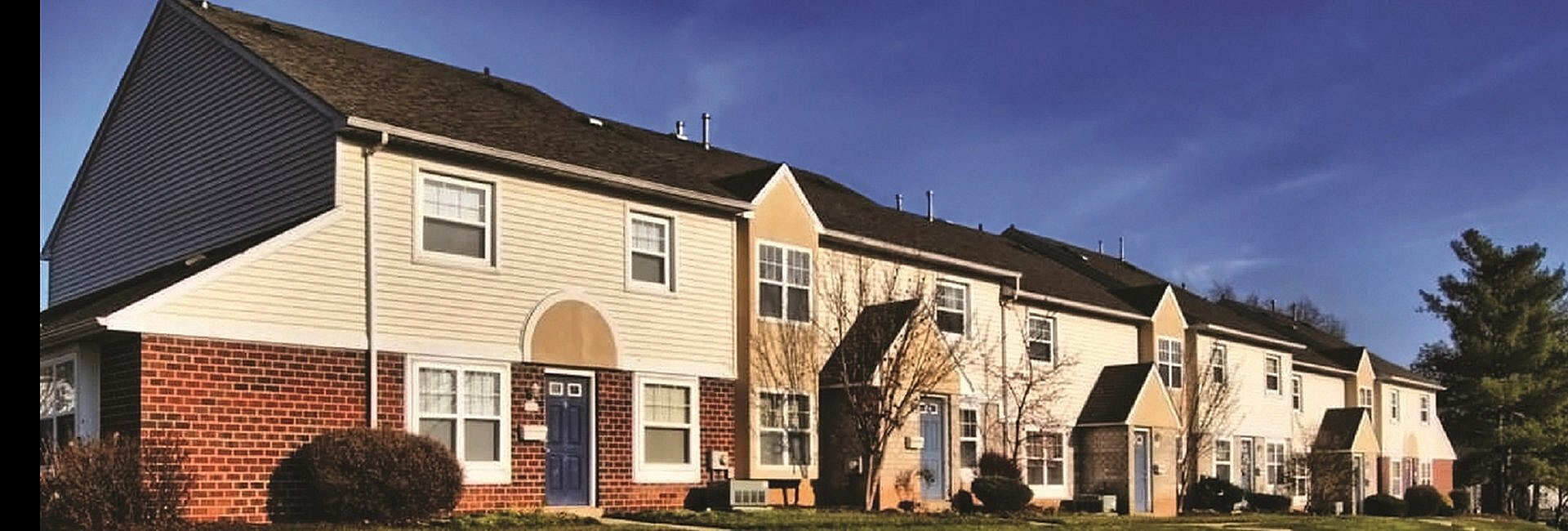 Charming townhouse row with brick and siding exteriors under a sunny blue sky, surrounded by lush greenery.