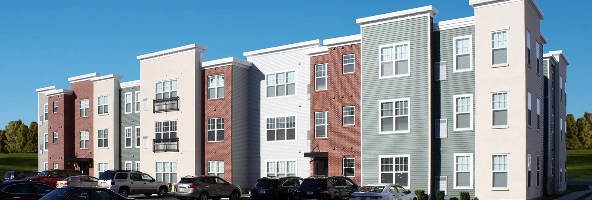 Modern apartment building with a vibrant mix of brick and siding finishes, ample parking, and lush greenery in the background under a clear blue sky.