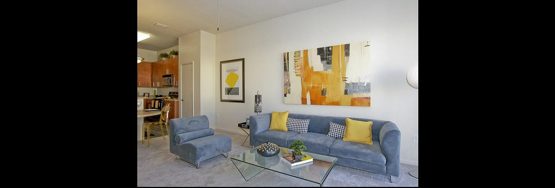 Modern living room featuring a chic blue sofa with vibrant yellow cushions, complemented by abstract wall art, leading to an open kitchen with sleek wooden cabinets.