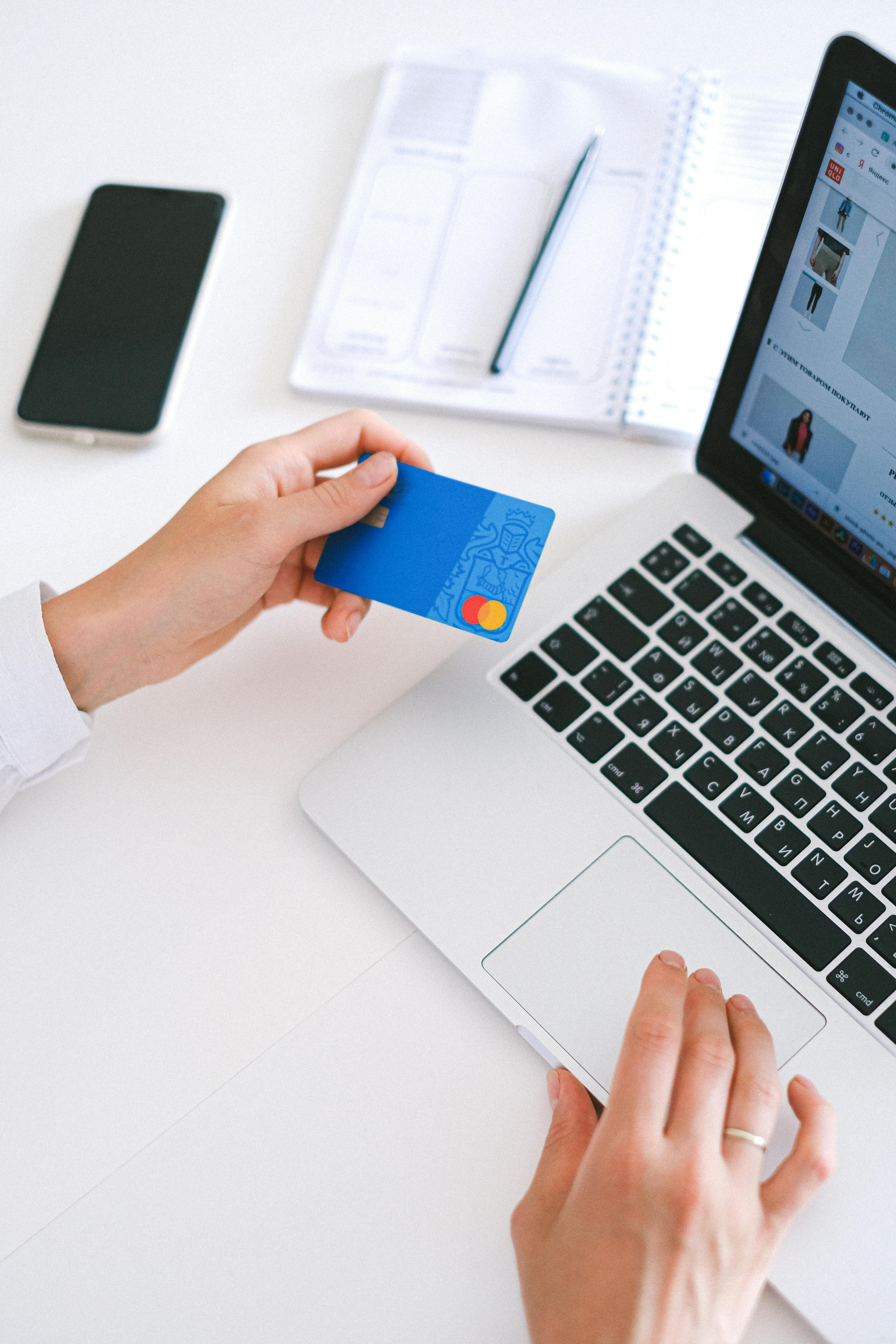 Person holding a blue credit card while using a laptop for online shopping, with a notebook and smartphone on a white desk.