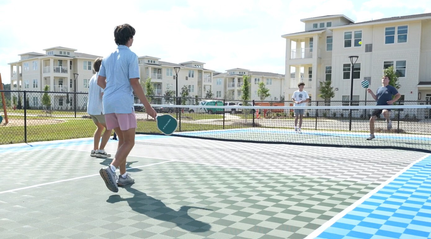 Modern apartment complex with vibrant pickleball match happening on a sunny day, showcasing outdoor recreation facilities and stylish residential buildings.