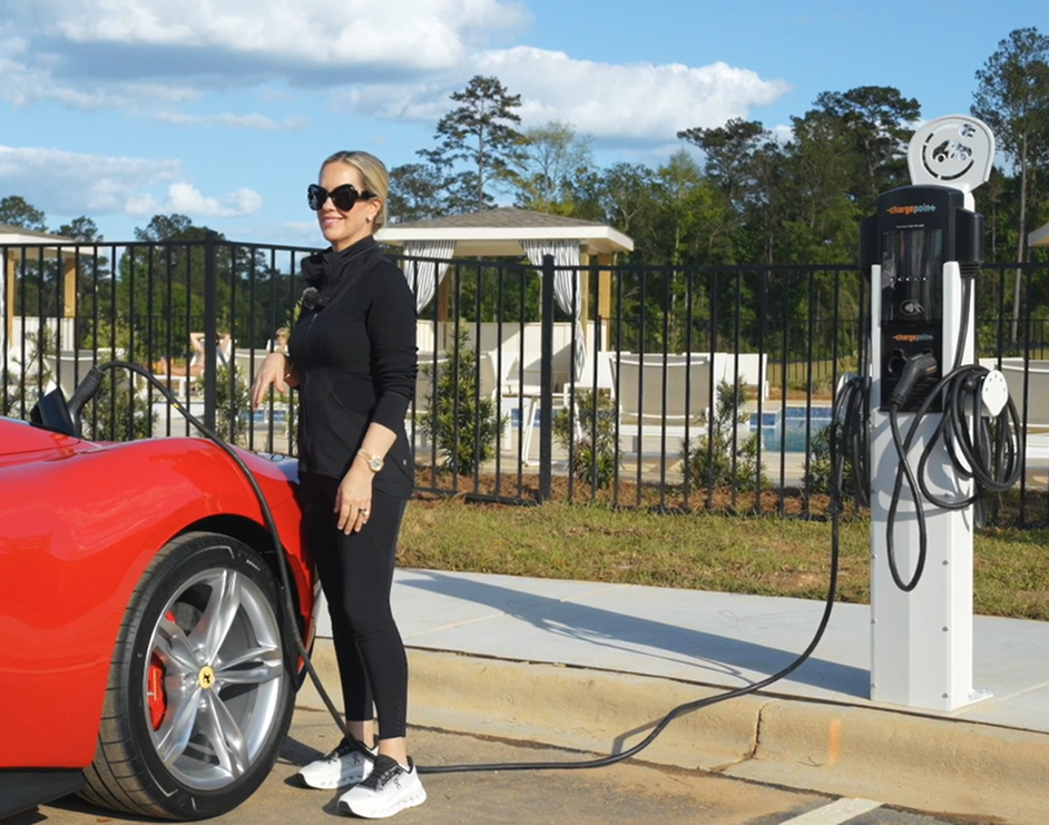 Chic woman in stylish sunglasses charging a vibrant red sports car at a modern electric vehicle station, set against a backdrop of lush greenery and a pool area.