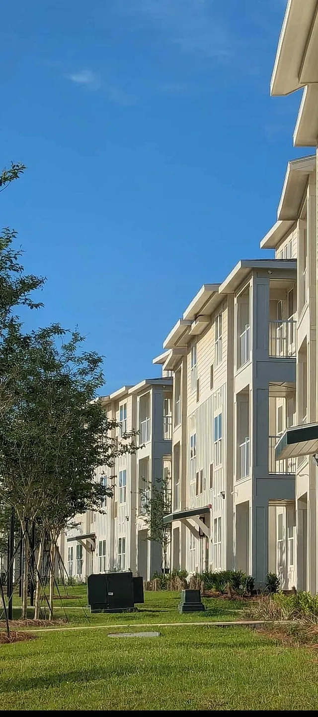 Modern apartment complex with sleek architecture and bright windows, surrounded by lush greenery under a clear blue sky.