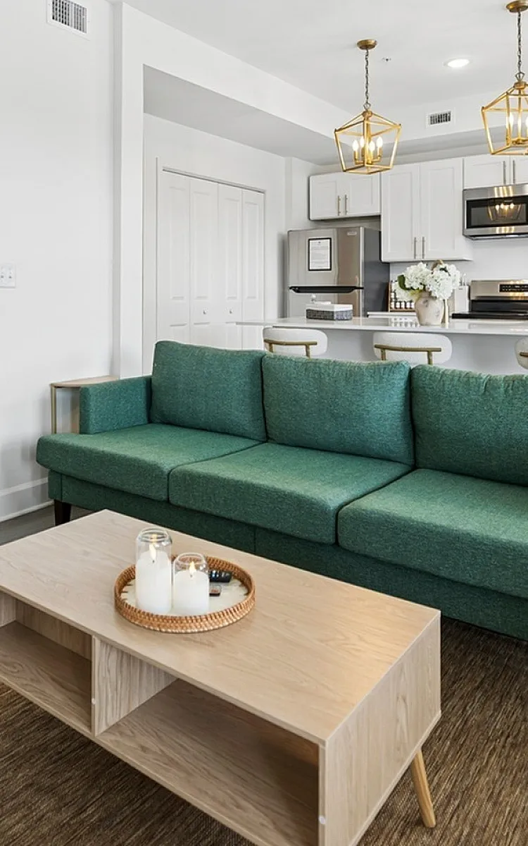 Modern apartment living room with a stylish green sofa, cozy armchair, and chic open kitchen featuring elegant lighting and white cabinetry.