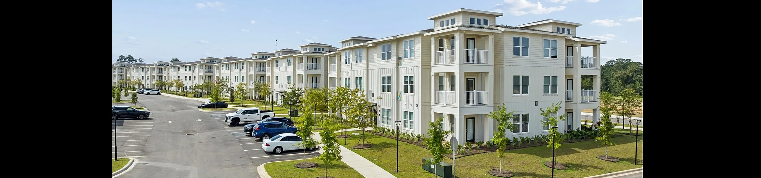 Modern apartment complex with elegant beige exterior, spacious balconies, surrounded by lush greenery, and ample parking space under a clear blue sky.