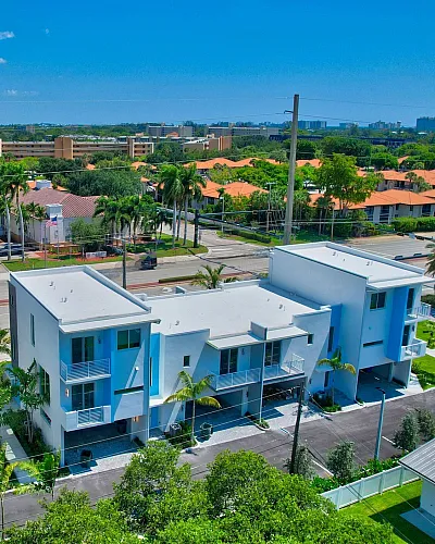 Modern townhouse complex with sleek design, surrounded by lush greenery and palm trees, offering vibrant community living under a clear blue sky.