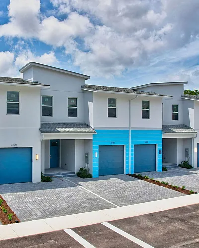 Modern townhouses with vibrant blue accents and spacious driveways under a bright, cloudy sky.