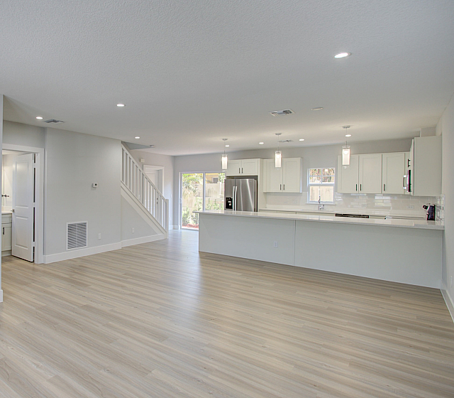 Modern open-concept living space with sleek hardwood floors, spacious kitchen featuring white cabinetry and stainless steel appliances, adjacent to a sunlit area with large windows.