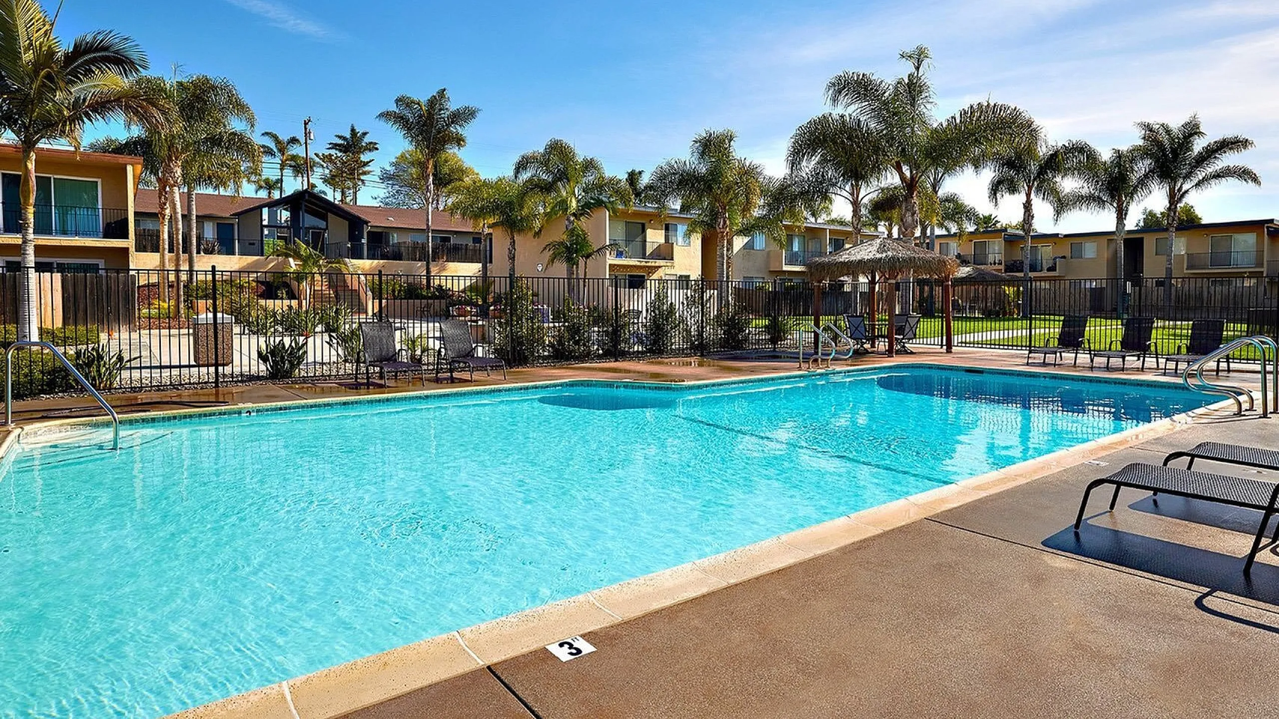 Luxurious apartment complex featuring a sparkling outdoor pool surrounded by lush palm trees and modern lounge chairs.