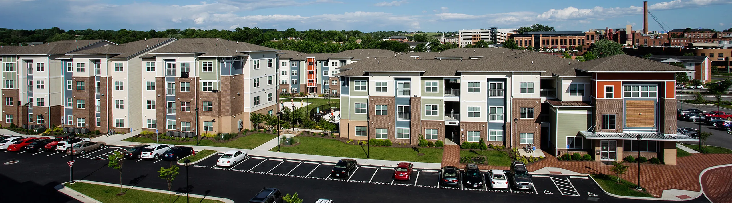 Modern apartment complex with vibrant exterior and ample parking, surrounded by lush greenery and cityscape in the background.