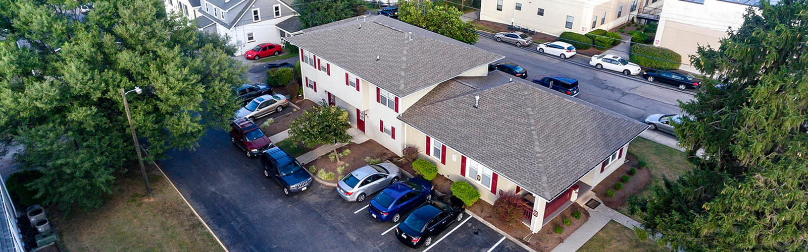 Aerial view of a charming residential complex with ample parking, surrounded by lush greenery and a serene neighborhood backdrop.