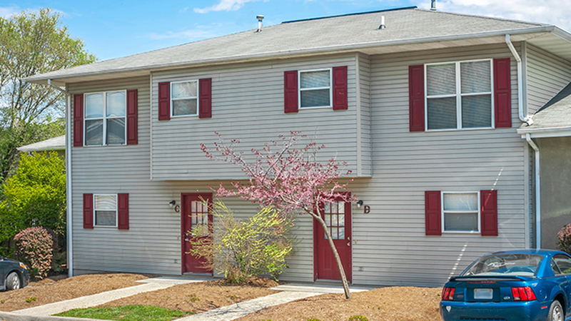Charming two-story apartment building with red shutters and doors, surrounded by neatly landscaped greenery under a bright blue sky.