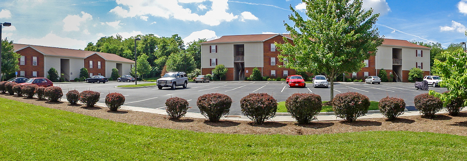 Charming apartment complex with lush landscaping, ample parking, and inviting exteriors on a sunny day.