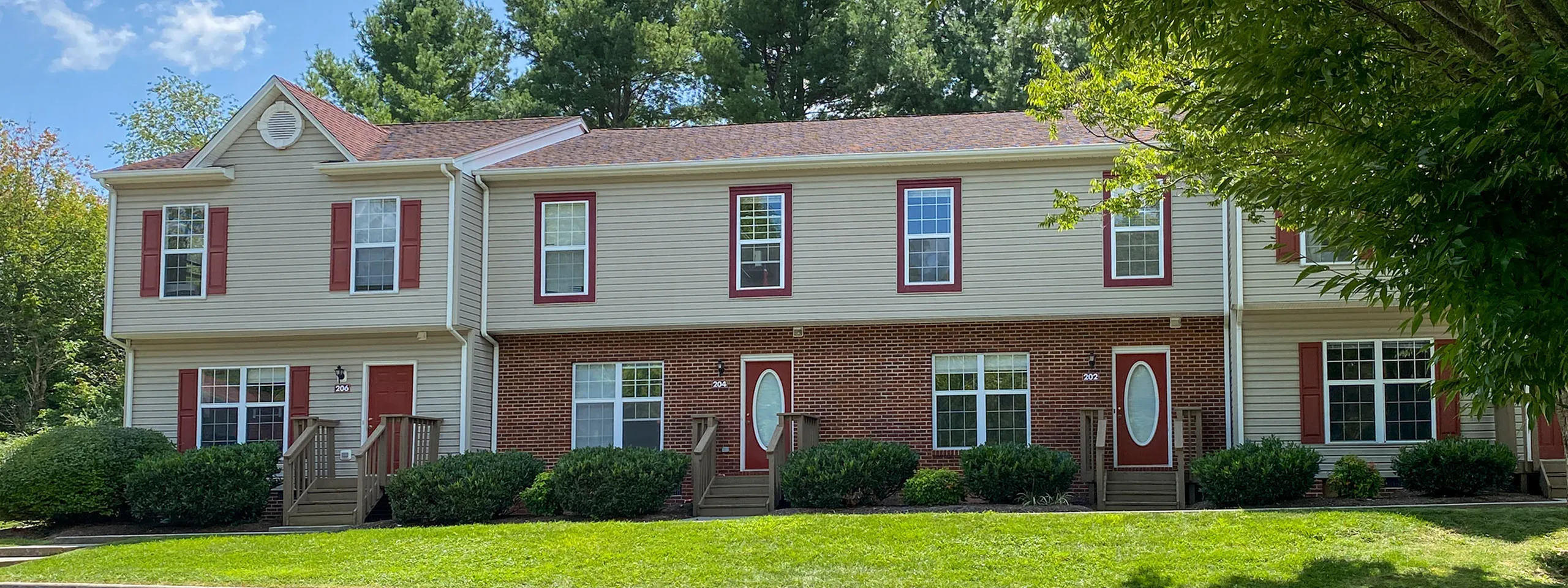 Charming townhouse with classic red shutters and inviting front porches, surrounded by lush greenery and mature trees.