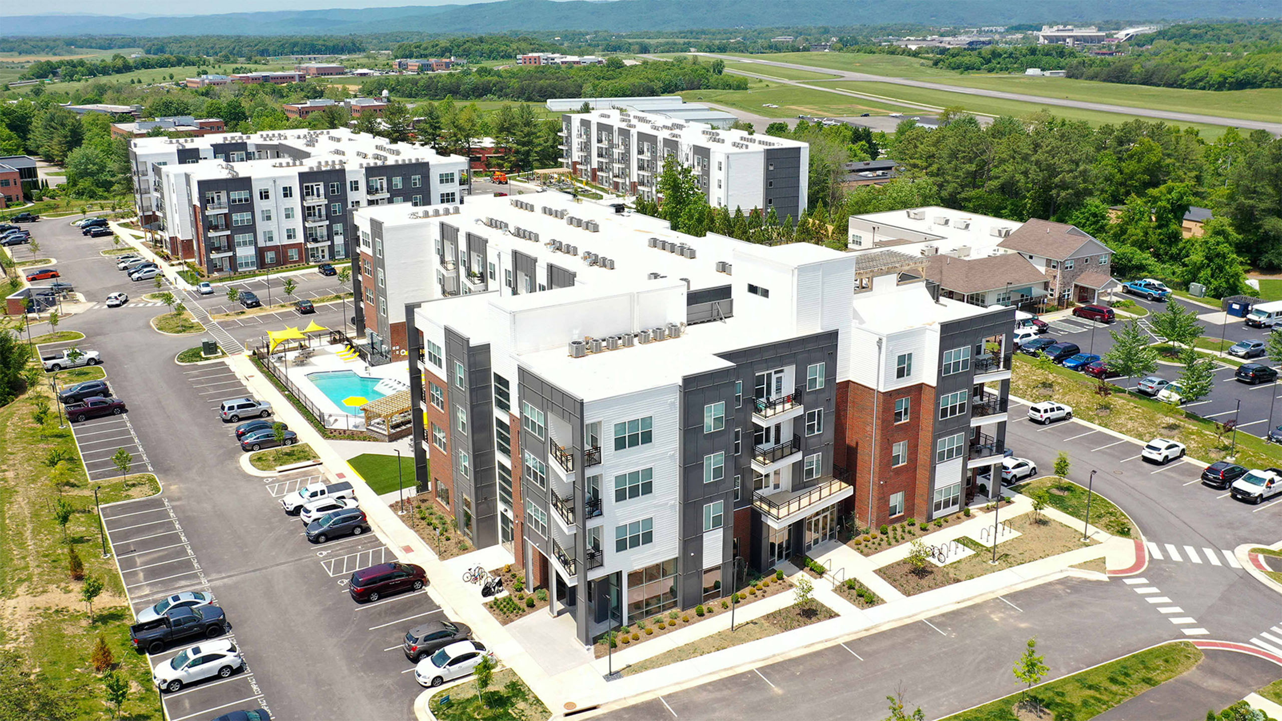 Aerial view of a modern apartment complex with spacious parking, featuring a central swimming pool and surrounded by lush greenery, providing an inviting and vibrant community feel.