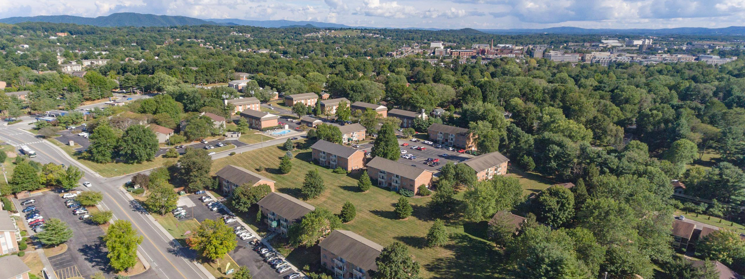 Aerial view of charming apartment community nestled in lush greenery. Features ample parking, paved roads, and scenic surroundings with distant mountain views.