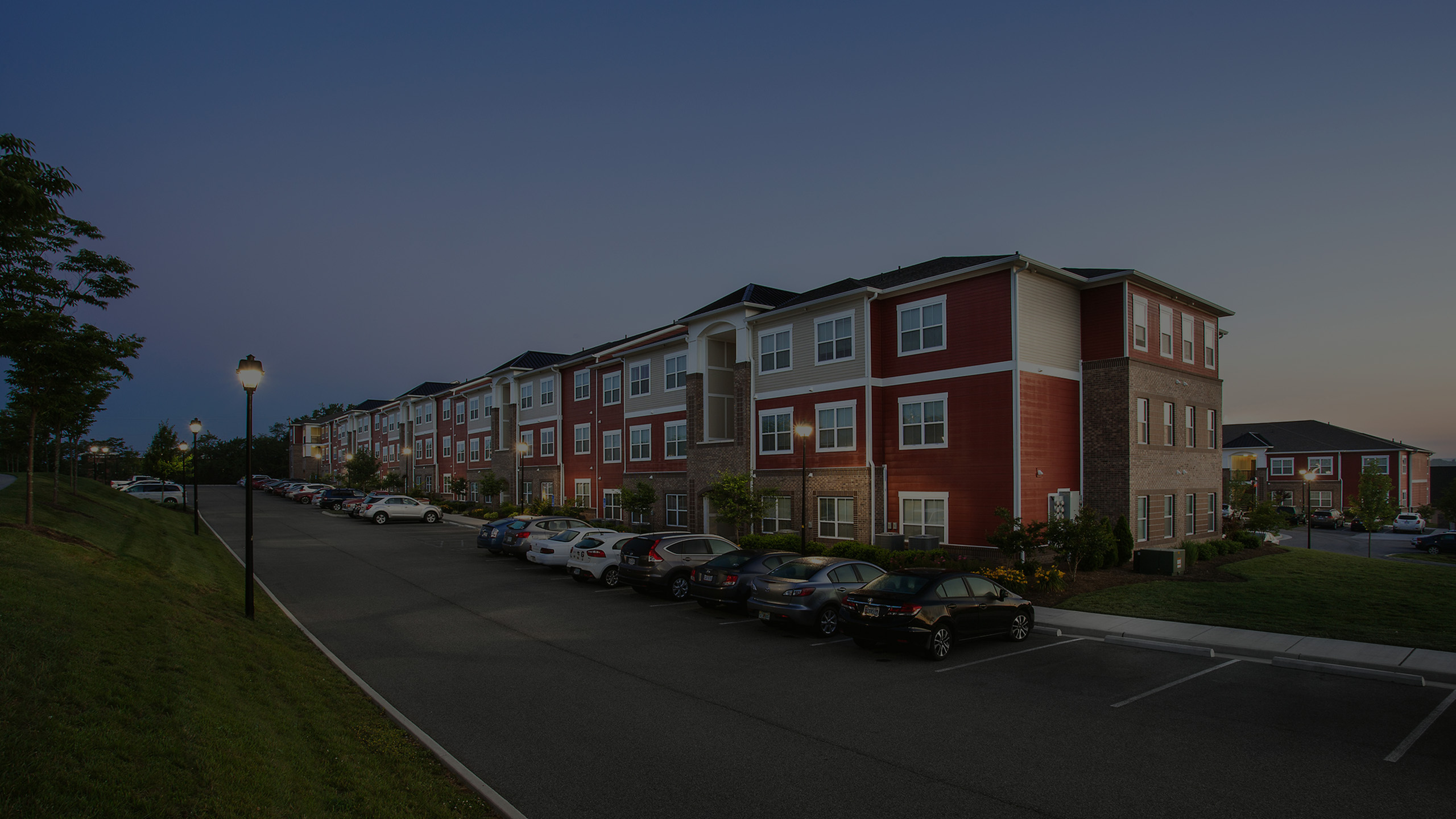 Charming apartment complex with vibrant red exteriors, beautifully landscaped surroundings, and ample parking, captured during a serene dusk setting.