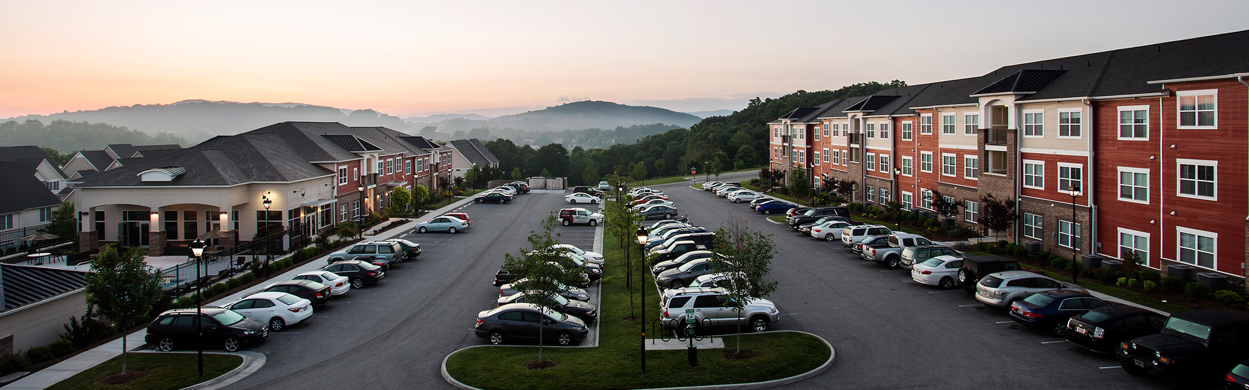 Scenic view of a modern apartment complex with ample parking, nestled in lush greenery against a beautiful mountain backdrop at sunrise.