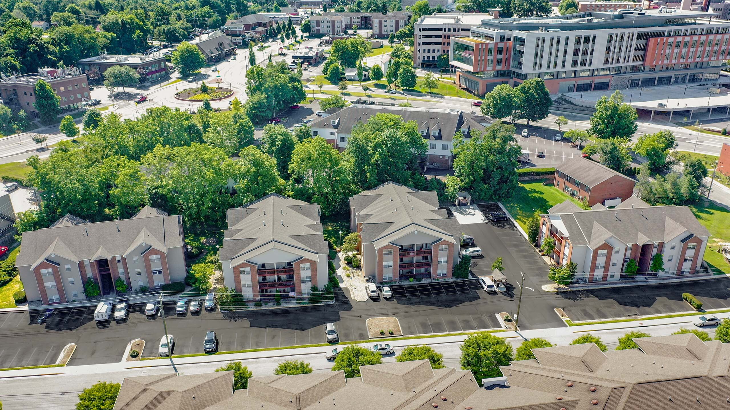 Aerial view of a charming apartment complex nestled among lush greenery, featuring spacious parking areas and a modern, urban setting.