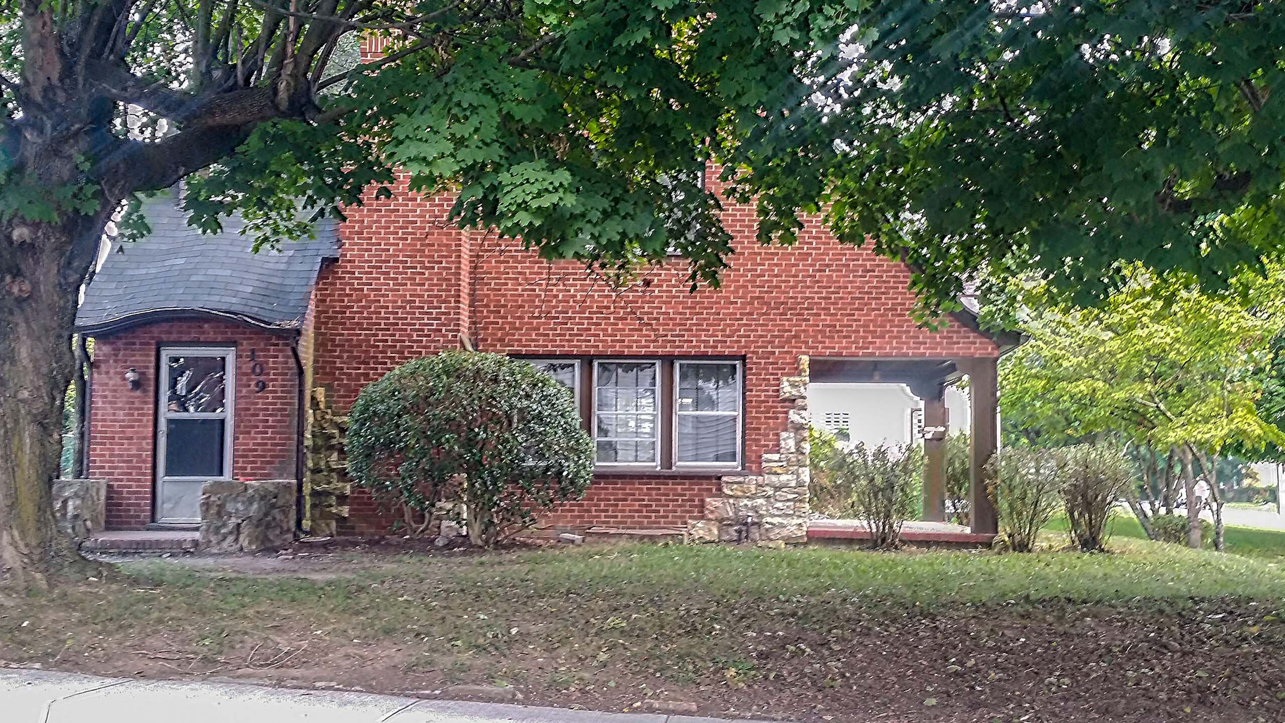 Charming brick house surrounded by lush greenery, featuring a quaint porch and scenic front yard.