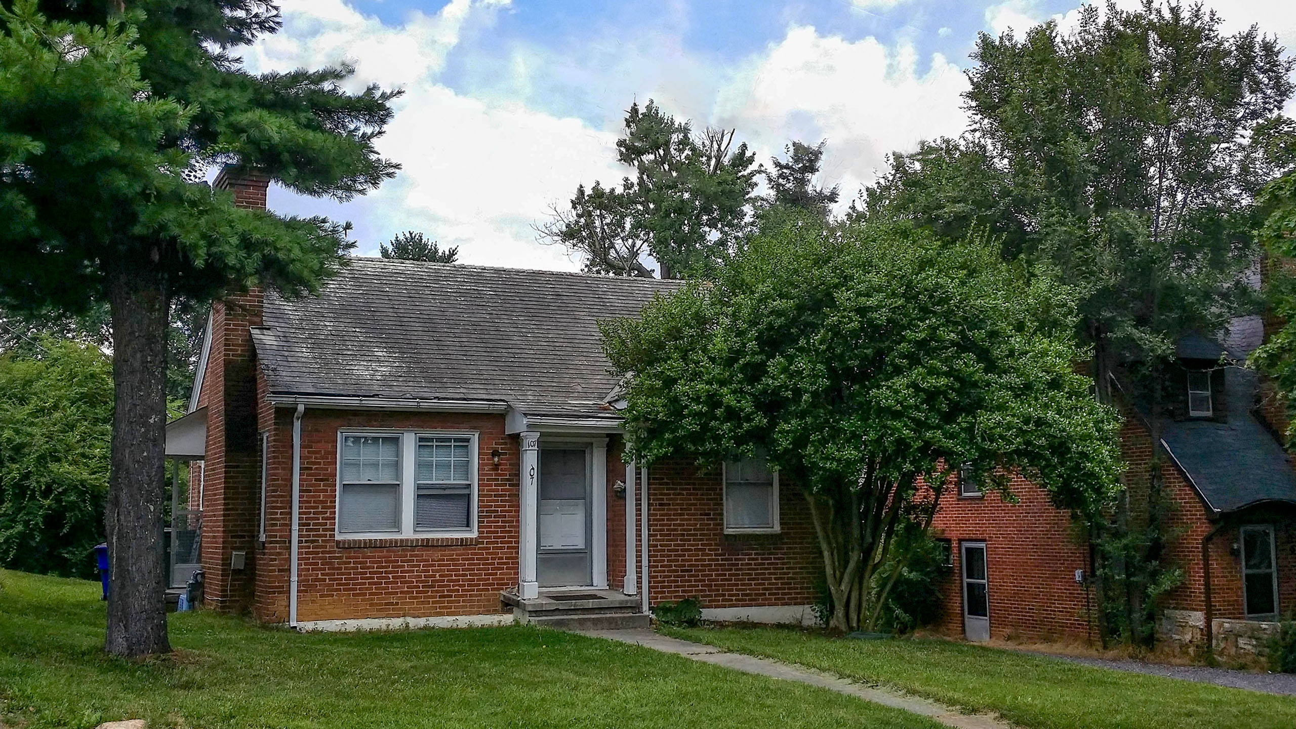 Charming red-brick house with a welcoming front porch, nestled among lush greenery and tall trees on a sunny day.