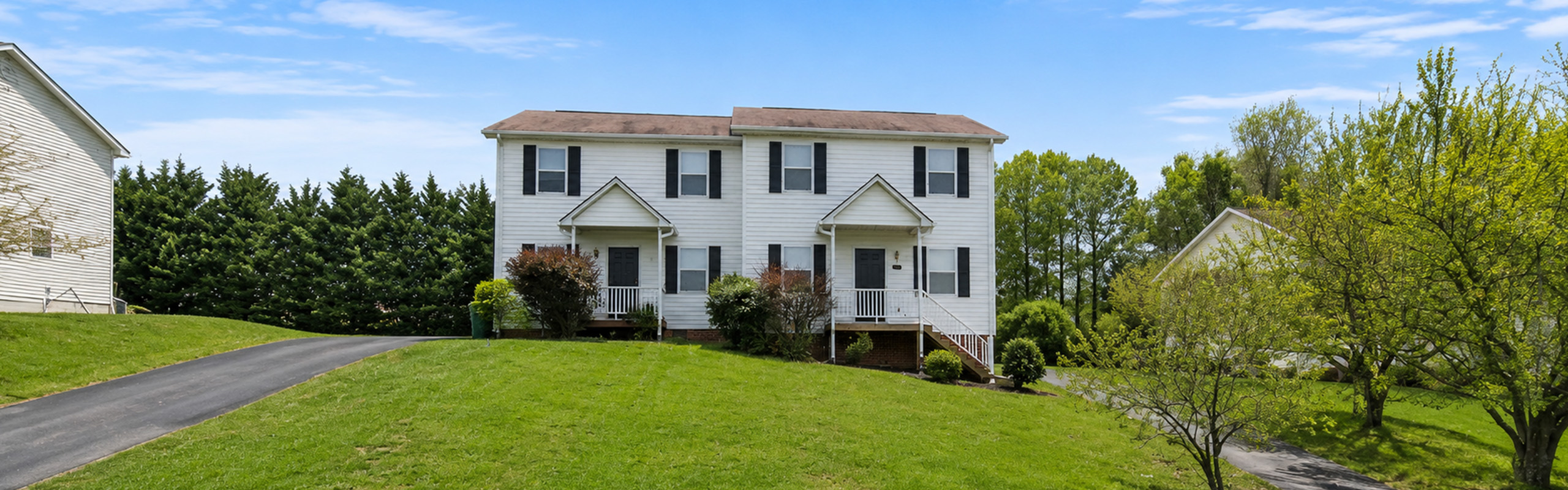 Charming two-story white house with black shutters, nestled in a lush green landscape and inviting driveway.