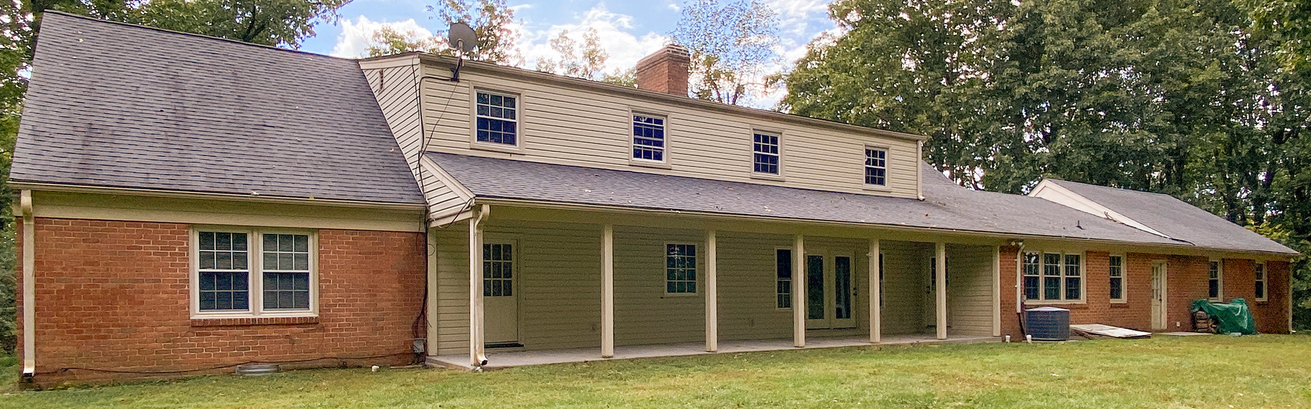 Charming countryside home with brick facade, elegant dormer windows, and a spacious covered porch surrounded by lush greenery.