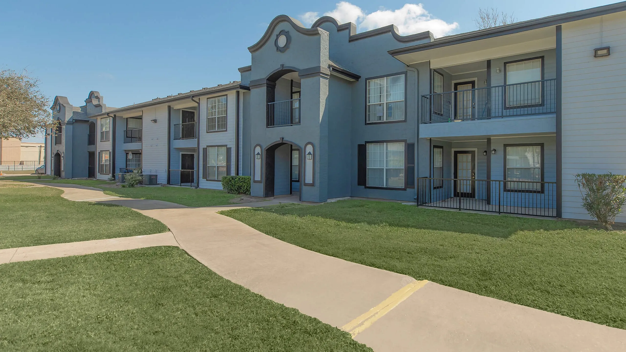 Charming two-story apartment complex with elegant arches, individual balconies, and lush green surroundings under a clear blue sky.