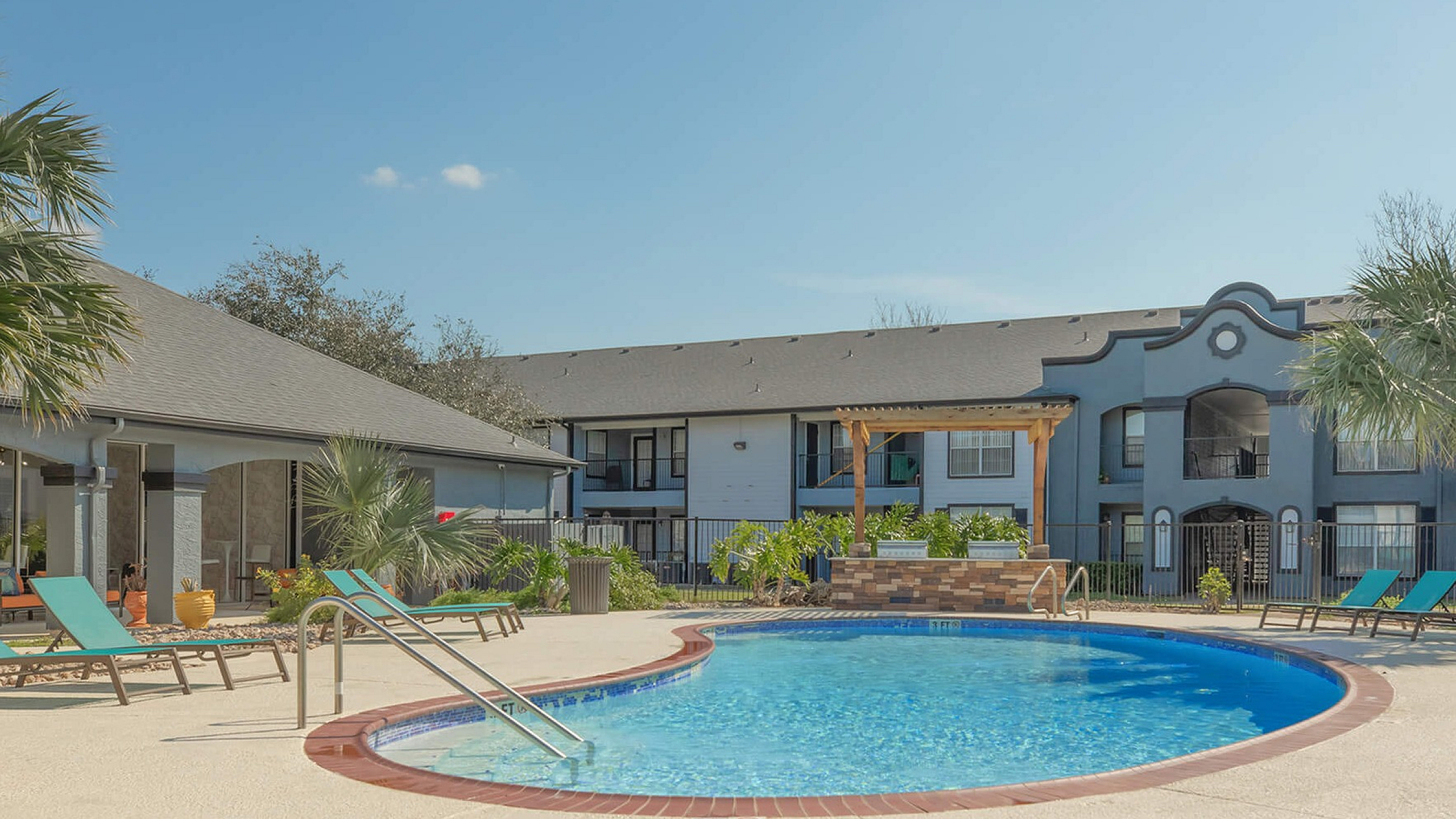 Charming apartment complex with a sunny swimming pool area, surrounded by lush palm trees and inviting lounge chairs.