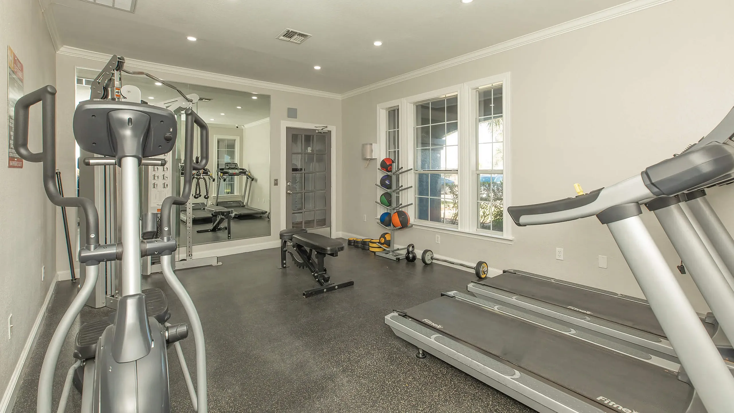 Modern fitness room featuring state-of-the-art treadmills, elliptical machine, and a variety of free weights with large windows providing natural light.