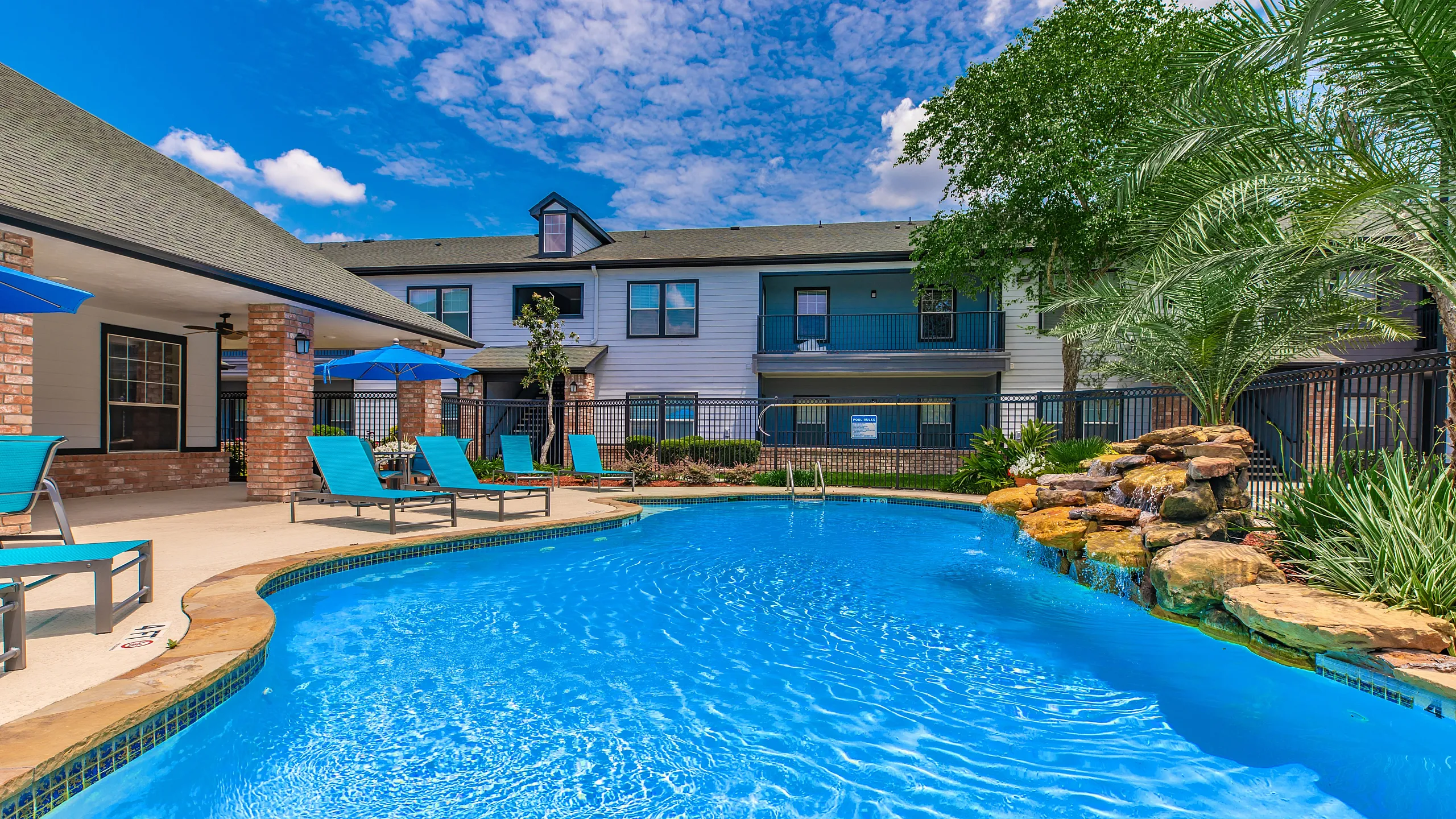 Inviting apartment complex with a pristine blue swimming pool, vibrant sun loungers, and lush greenery, perfect for relaxation and leisure.