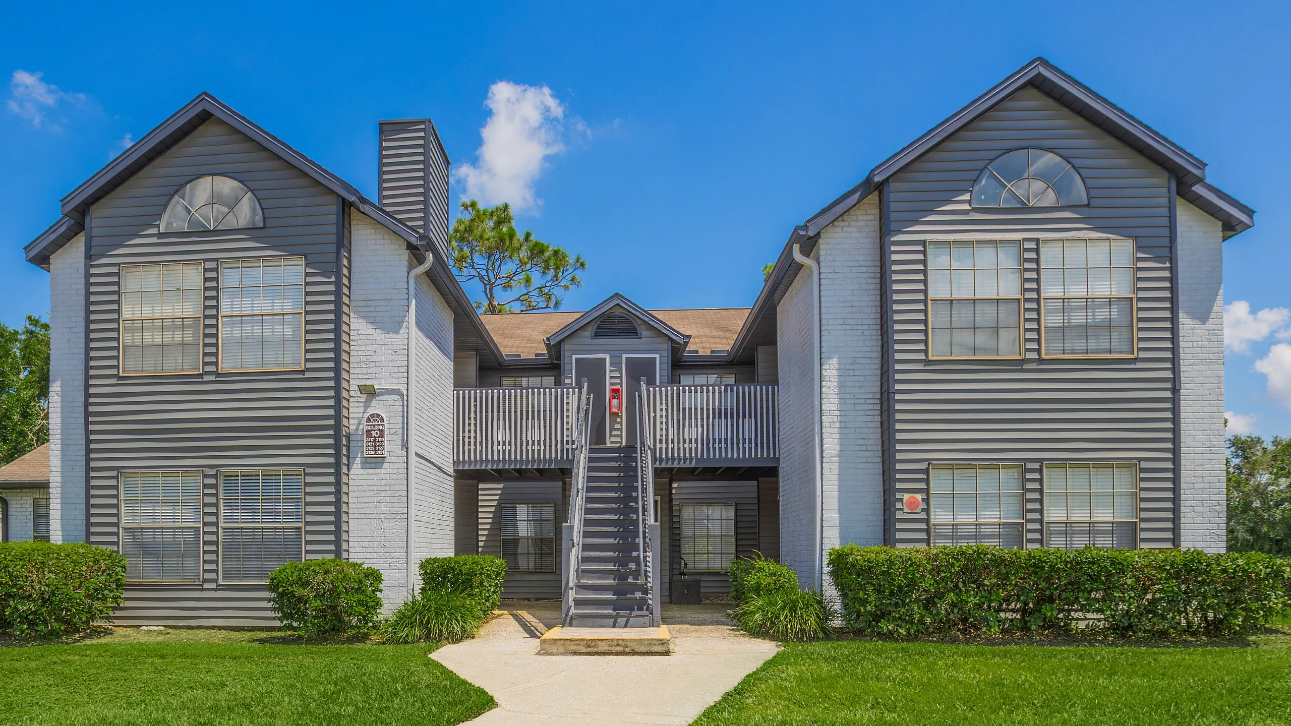 Charming two-story apartment building with gray siding, lush green lawn, and picturesque walkway on a sunny day.