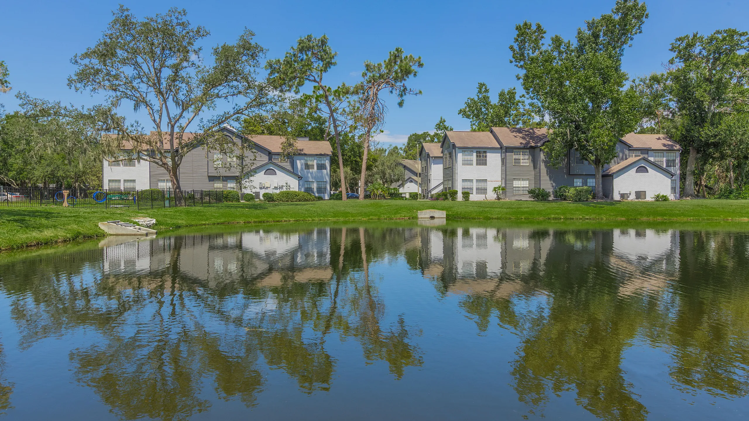 Charming apartment buildings reflected in a serene pond, surrounded by lush greenery and clear blue skies.