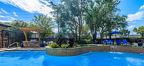 Inviting outdoor pool area with a serene blue pool, elegant stone pergola, lush greenery, and comfortable lounge chairs under vibrant blue umbrellas.