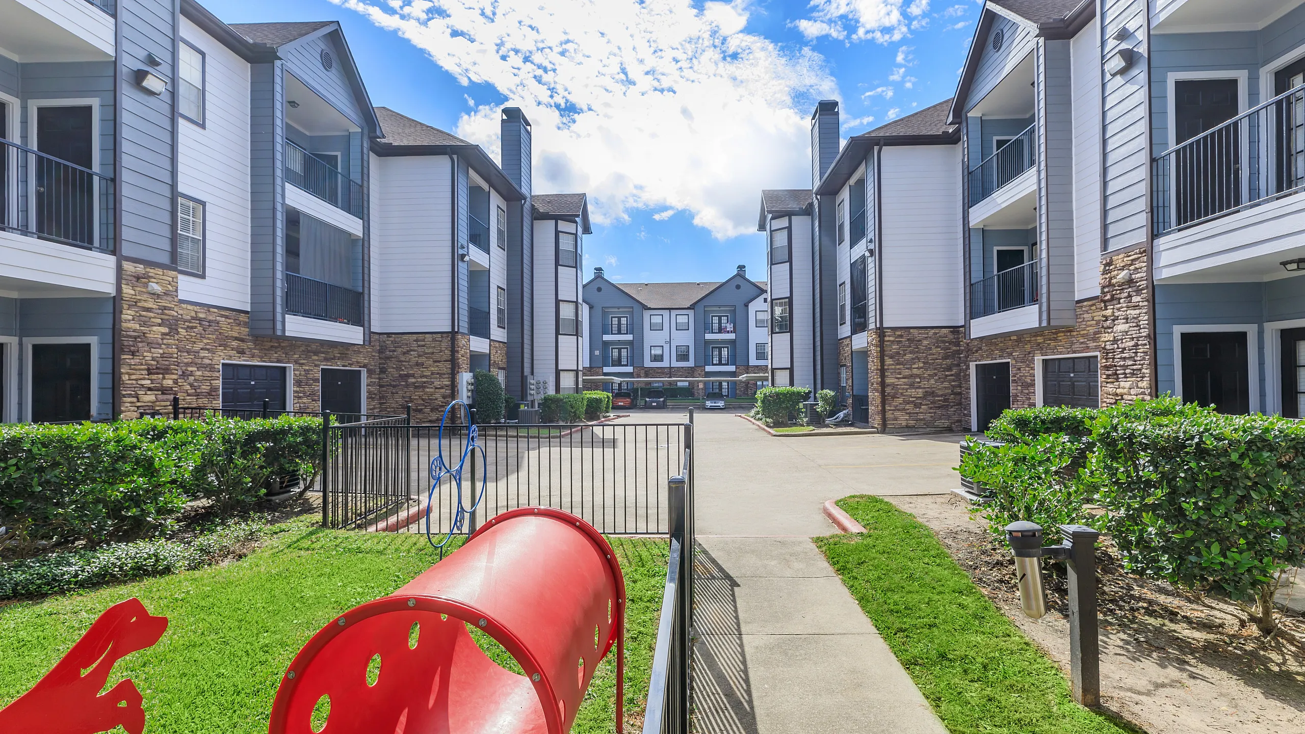 Modern apartment complex with elegant design, lush green landscaping, and family-friendly play area, set against a bright blue sky.