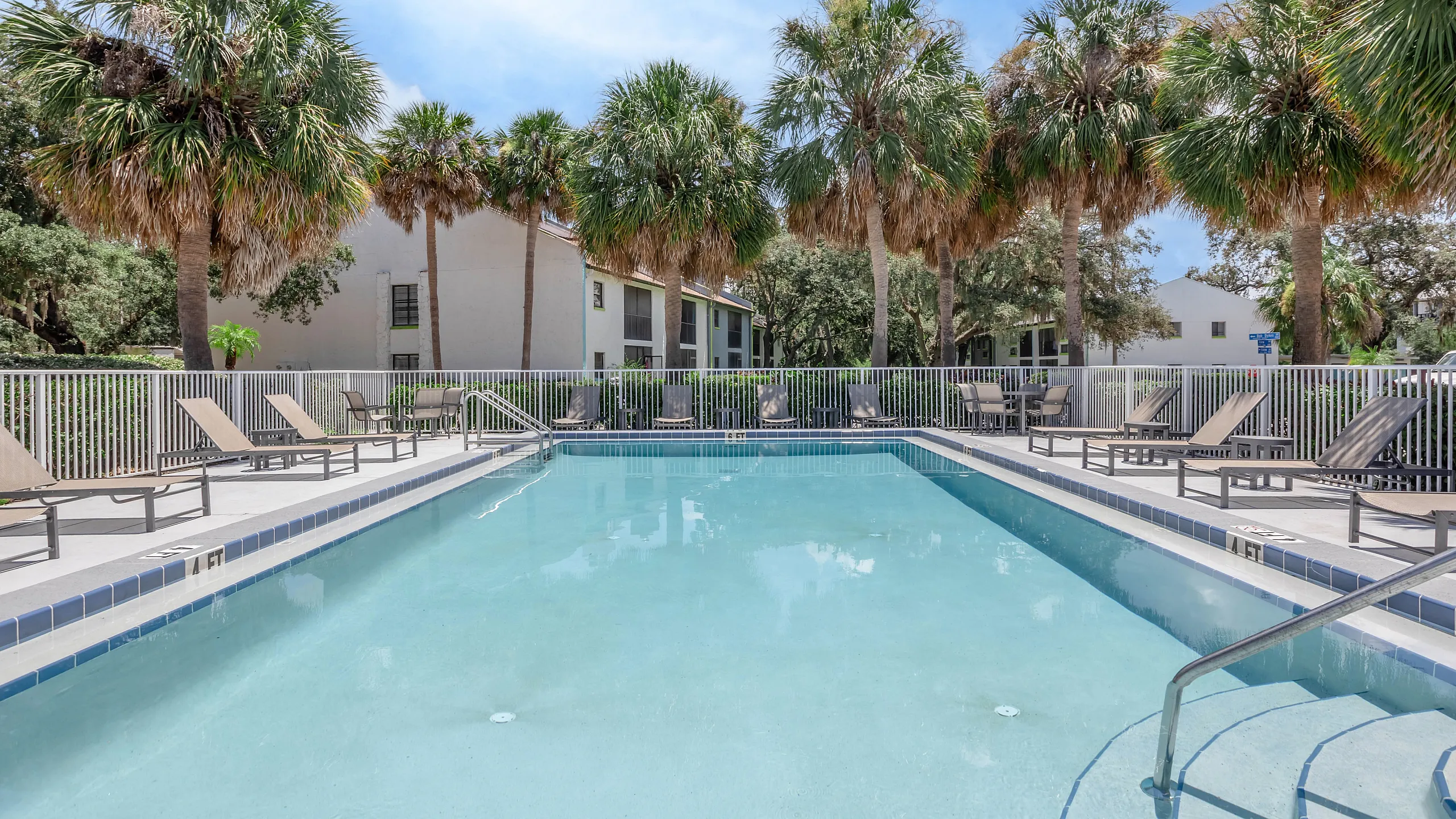 Luxurious apartment community pool area featuring a sparkling swimming pool surrounded by comfortable lounge chairs and lush palm trees, offering a tropical oasis for relaxation.
